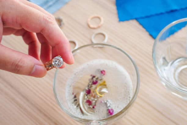 A hand dips a ring into a glass bowl with soapy water containing various jewelry pieces on a wooden surface.