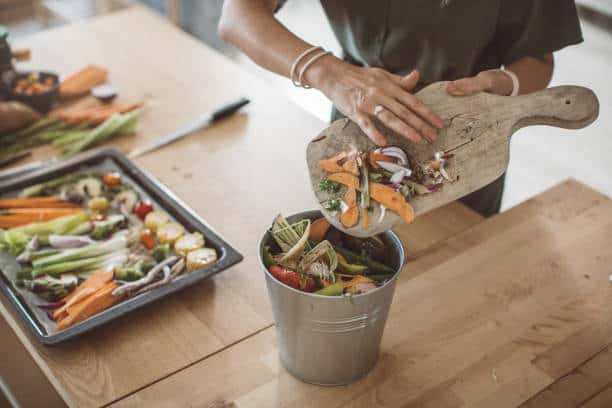 A person is scraping vegetable scraps from a cutting board into a metal compost bin on a wooden table, with a tray of chopped vegetables in the background.