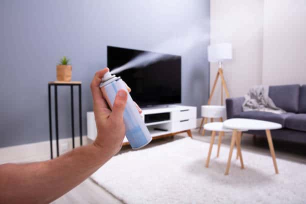 A person is spraying an aerosol can in a living room with a gray couch, white round tables, a TV, and a lamp in the background.