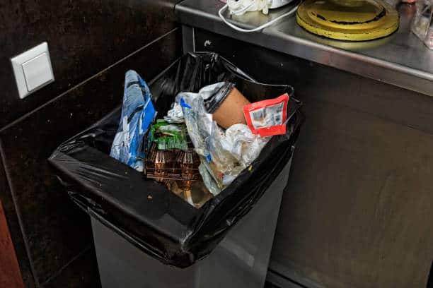 A trash bin filled with various types of waste, including plastic packaging, paper, and other discarded items, located in a kitchen area.