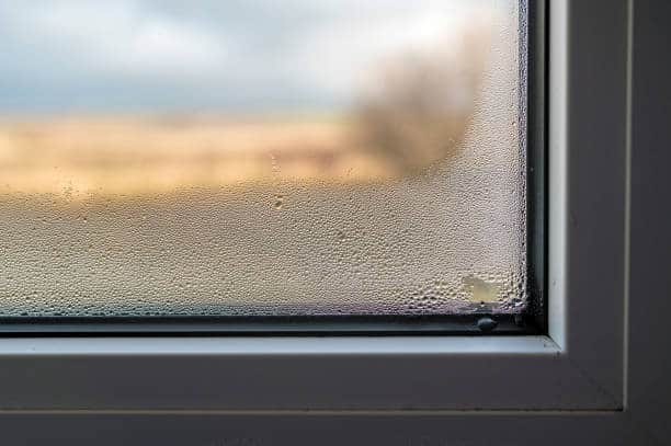 Close-up of a window with condensation on the pane, showing water droplets forming on the glass. The view outside is blurred.