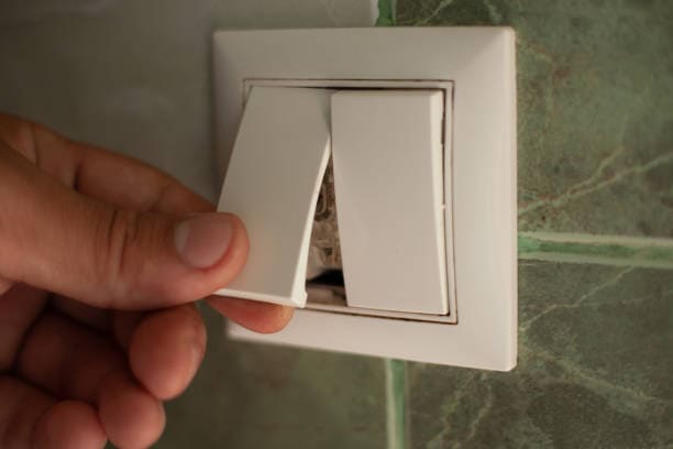 A hand is fixing a broken white light switch on a green tiled wall.