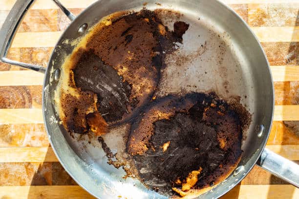 A stainless steel pan with heavily burnt remnants of food on its surface, placed on a wooden countertop.