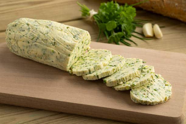 Sliced herb butter log on a wooden cutting board, surrounded by fresh herbs and garlic cloves.