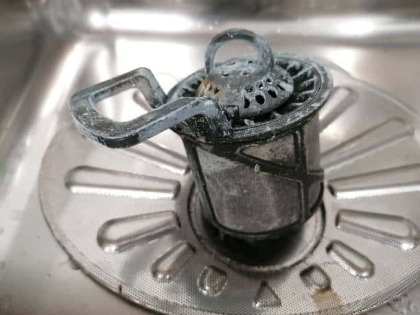 A close-up of a dirty, metal kitchen sink strainer with accumulated grime and debris, placed in the sink's drain area.