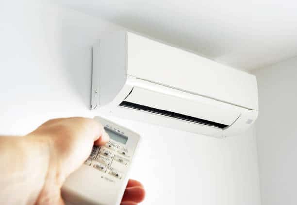 A hand holds a remote control pointed at a wall-mounted air conditioning unit.