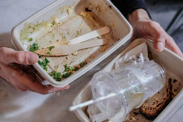Two hands hold empty food containers: one with wooden cutlery and food scraps, and the other with a used plastic cup, napkin, and leftover crusts.