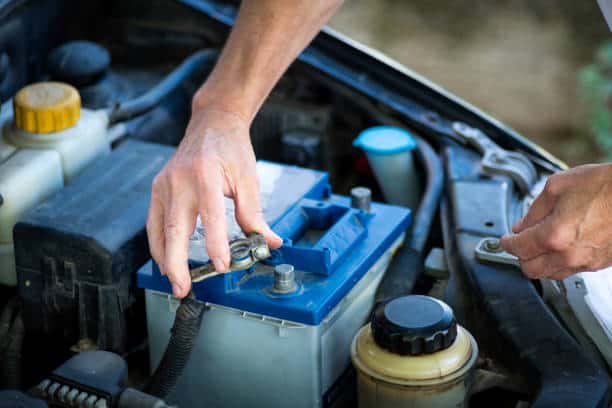 Hands adjusting cables on a blue car battery under the open hood.