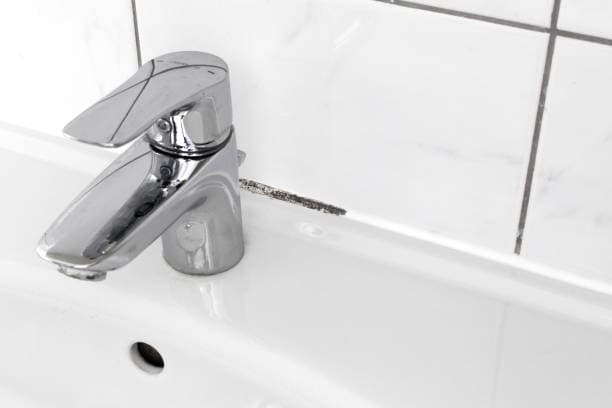A close-up of a silver faucet attached to a white bathroom sink with a small patch of black mold on the tile grout behind it.