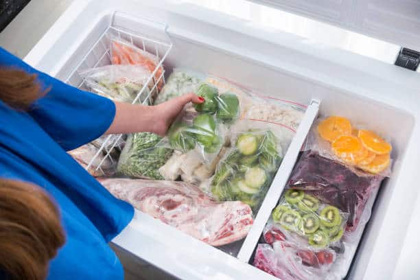 Person placing green bell peppers in a chest freezer filled with various frozen foods, including vegetables and fruit slices.