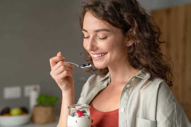 A woman with curly hair smiles while eating yogurt with berries from a jar using a spoon.