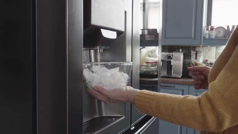 A person fills a glass bowl with ice from a refrigerator dispenser in a kitchen.