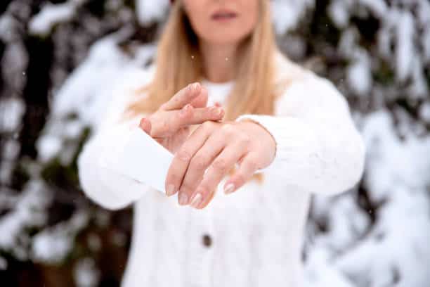 Person in a white sweater applies cream to their hands outdoors with a snowy background.