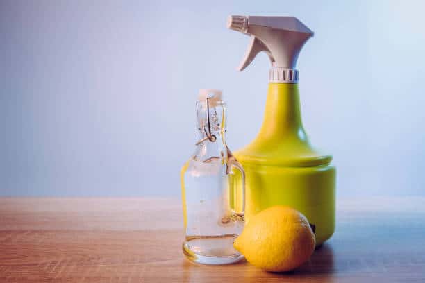 A glass bottle, a yellow spray bottle, and a lemon are placed on a wooden surface against a light blue background.