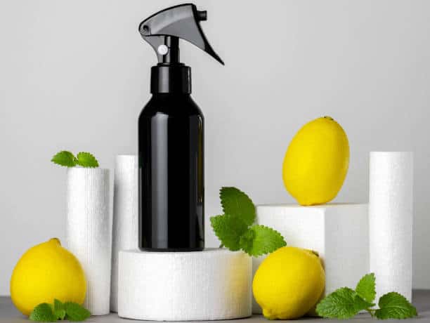 A black spray bottle on white cylindrical stands, surrounded by three lemons and green leaves against a gray background.