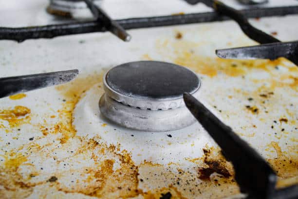 Close-up of a dirty stovetop burner with grime and food stains on a white surface, surrounded by black grates.