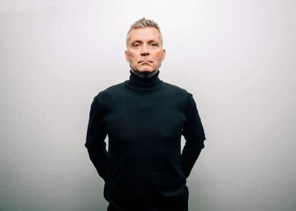 A man in a black turtleneck stands against a plain light gray background with his hands behind his back.