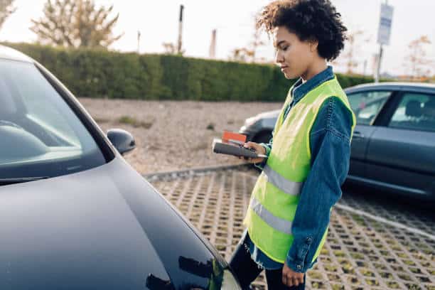 A person in a yellow safety vest writes a ticket for a parked car in an outdoor parking lot.
