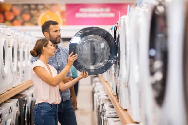 A man and woman examine a washing machine in an appliance store, standing in an aisle lined with machines.
