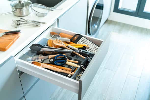 Open kitchen drawer with various utensils, including scissors, tongs, and a whisk, next to a countertop with a cutting board and cutlery.