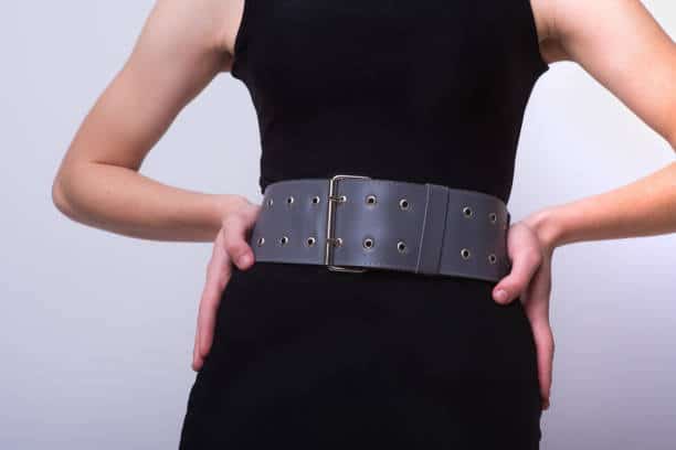 A person wearing a black dress and a wide gray belt stands with hands on hips.
