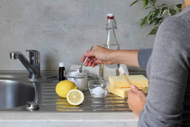 Person preparing cleaning solution with baking soda, lemon, vinegar, a bottle, sponges, and essential oil on a kitchen sink.