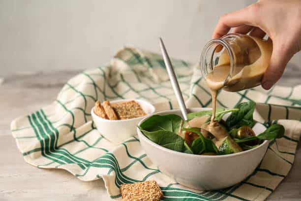 Hand pouring dressing over a spinach salad in a white bowl, with crackers on the side. A green and white checkered cloth is underneath.