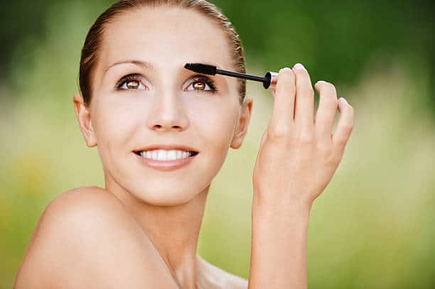 A person smiles while applying mascara with a brush outdoors, against a blurred natural background.