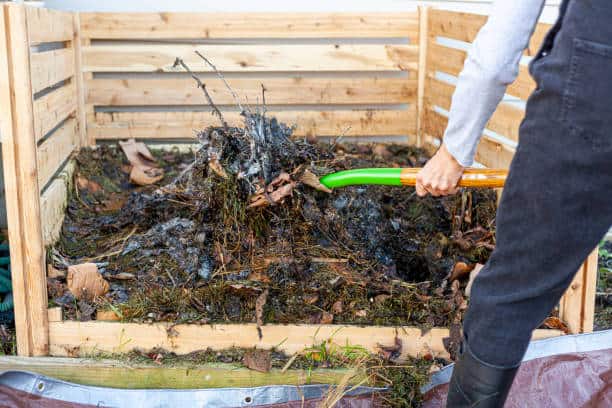 Person turning compost with a green-handled tool in a wooden compost bin filled with leaves, grass, and plant material.
