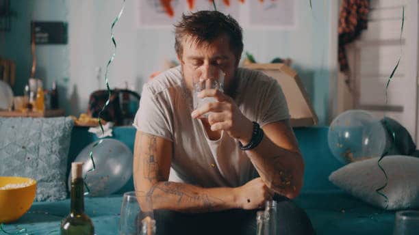A man with tattoos sits on a blue sofa, drinking from a glass. Balloons and streamers are around him, with an open bottle and bowl of snacks nearby.