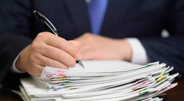 A person in a suit holds a pen, poised to write on a stack of papers clipped together with colorful paper clips.