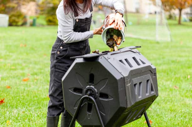 A person in black overalls pours kitchen scraps from a bucket into a black outdoor compost bin situated on a grassy lawn.