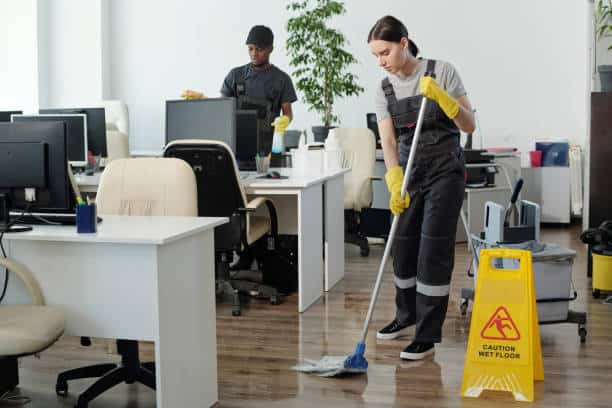 Two office cleaners wearing uniforms are mopping the floor. A "Caution Wet Floor" sign is visible. Desks and computers are in the background.