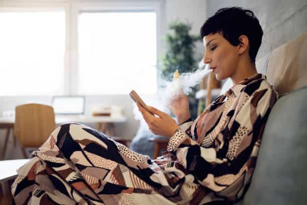 A person in patterned clothing sits on a sofa, using a phone with one hand while holding a vape and exhaling vapor with the other.