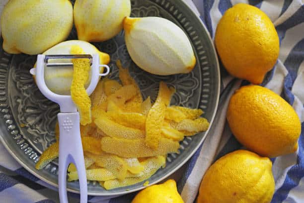 Peeled and unpeeled lemons on a plate with a peeler and lemon zest on a striped cloth.