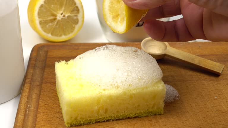 A person squeezes lemon juice onto a yellow sponge with soapy foam on a wooden board. A half lemon and wooden spoon are in the background.