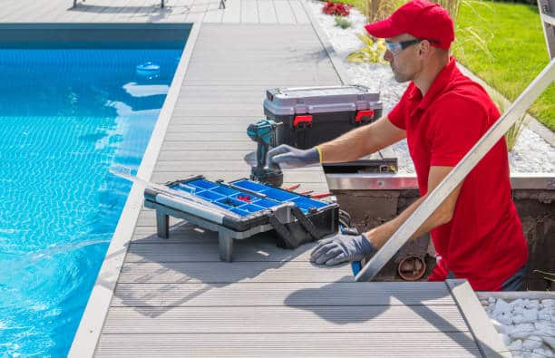 A worker in a red uniform and cap performs maintenance next to a swimming pool, using tools from a box.