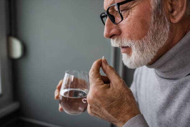 An older man with glasses holds a pill in one hand and a glass of water in the other.