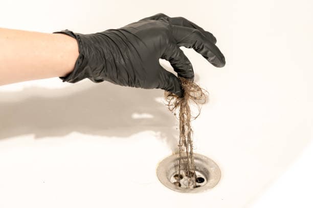 A person wearing black gloves removes tangled hair from a bathtub drain.