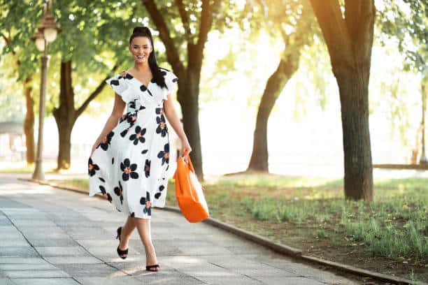 A woman in a white floral dress carries an orange bag while walking on a tree-lined path in sunlight.