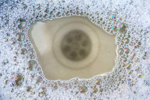 Close-up of a sink drain with soapy water and bubbles surrounding it. The drain's circular opening is visible in the center.