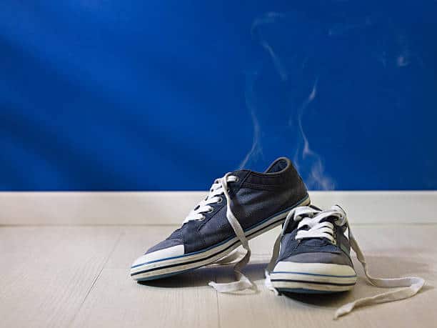 A pair of black canvas sneakers with white laces on a wooden floor against a blue wall. One shoe emits visible smoke.