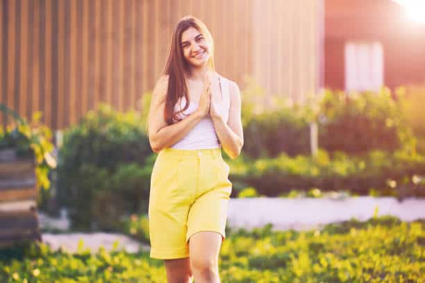 A woman with long hair is smiling and standing outdoors, wearing a white top and yellow shorts. The background features greenery and a wooden structure.