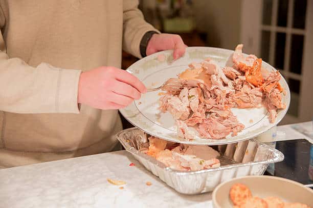 Person transferring leftover turkey from a serving plate to an aluminum tray in a kitchen.