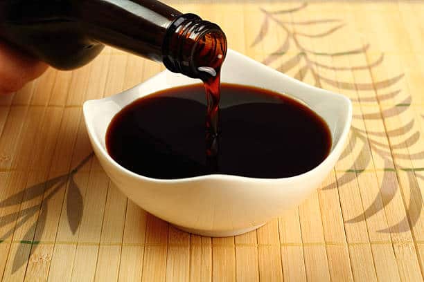 Soy sauce being poured into a small, white bowl on a bamboo mat.