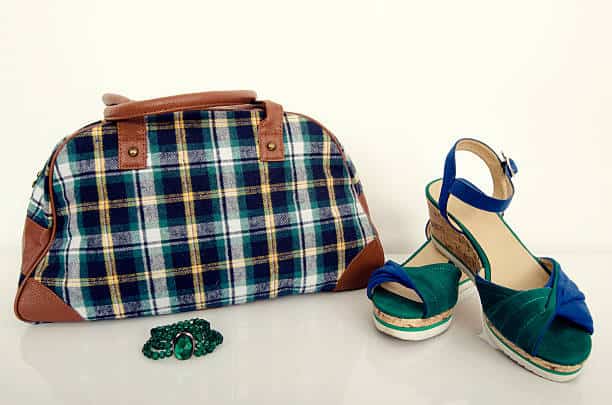A plaid handbag, a pair of green and blue wedge sandals, and a green beaded bracelet are displayed on a white surface.