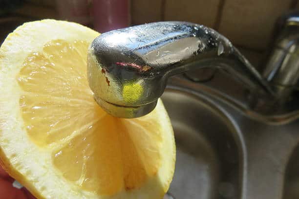 A halved lemon is held against a kitchen faucet over a sink.