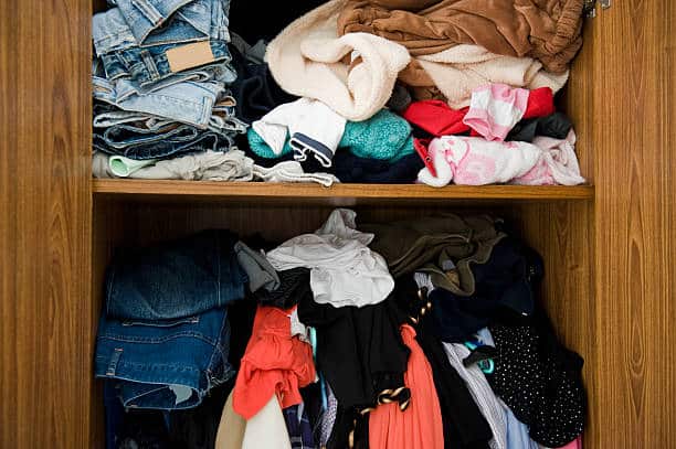 A cluttered wardrobe with disorganized stacks of clothes, including jeans, shirts, and various other clothing items, on two wooden shelves.