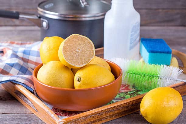 A bowl of lemons with a halved lemon on top, surrounded by a scrub brush, sponge, and bottle, on a wooden tray near a checkered cloth and pot.