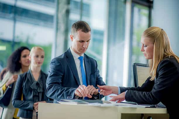 A man in a suit checks in at a reception desk. A woman behind the desk assists him. Two people wait in line in the background.